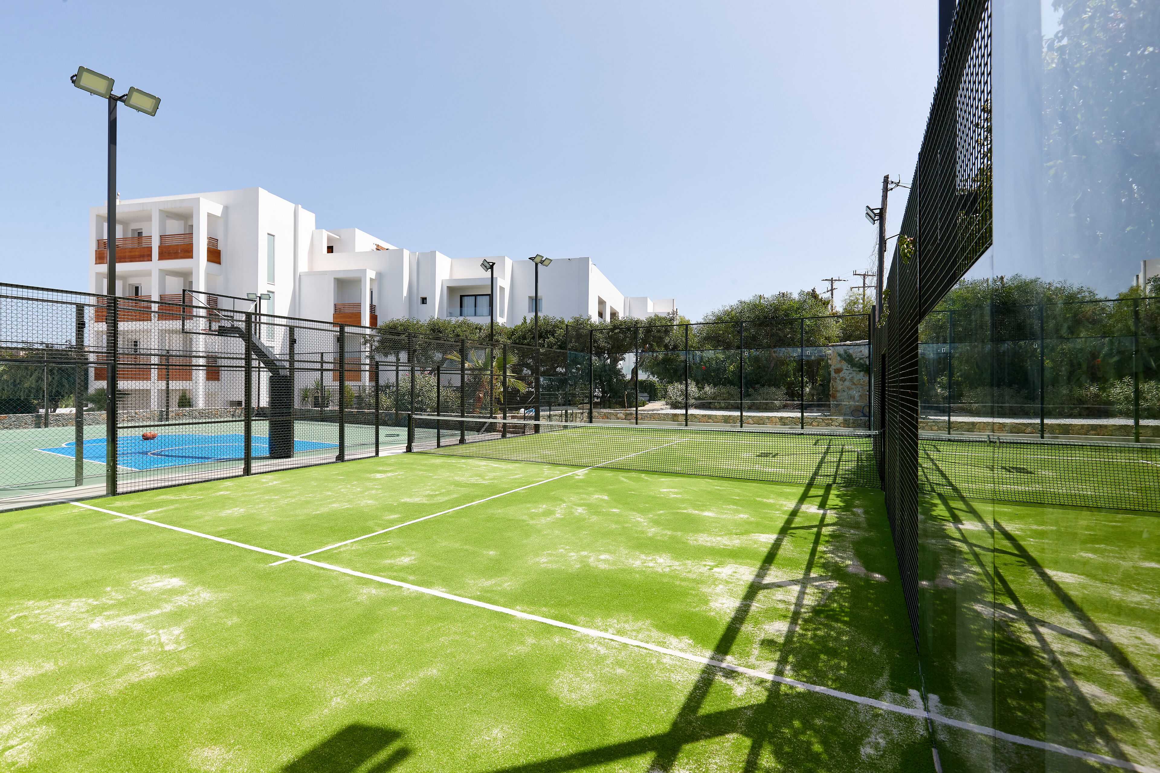 Een tennisbaan met een net, omringd door een hek, en een gebouw met balkons op de achtergrond.