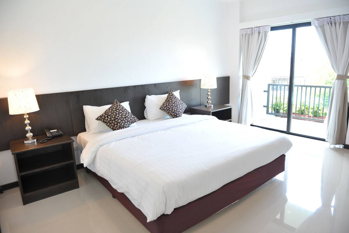 Habitación de hotel con una cama grande, dos mesitas de noche con lámparas, un teléfono y vistas al exterior desde un balcón.