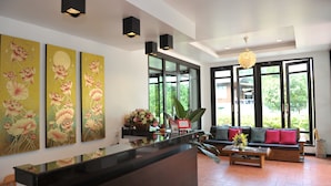 Un área de recepción con un mostrador negro, tres cuadros florales, un sofá con cojines rojos y ventanas amplias con vistas al verdor.
