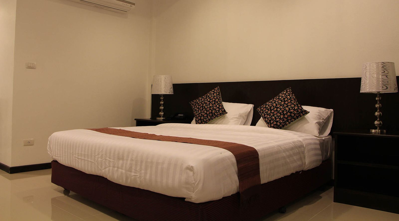 Una cama bien hecha con una colcha blanca y marrón, dos almohadones decorativos y dos lámparas de noche a cada lado.