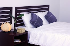 A neatly made bed with white bedding and dark blue pillows, a bedside table with a lamp and a small decorative item, and a dark wooden headboard. - Niisaii Apartment (Phnom Penh)