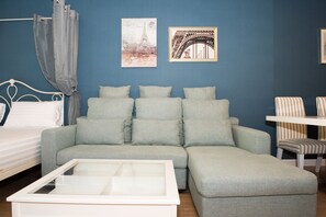 Quarto com sofá cinzento, cama branca, mesa de centro de vidro e duas imagens emolduradas na parede.