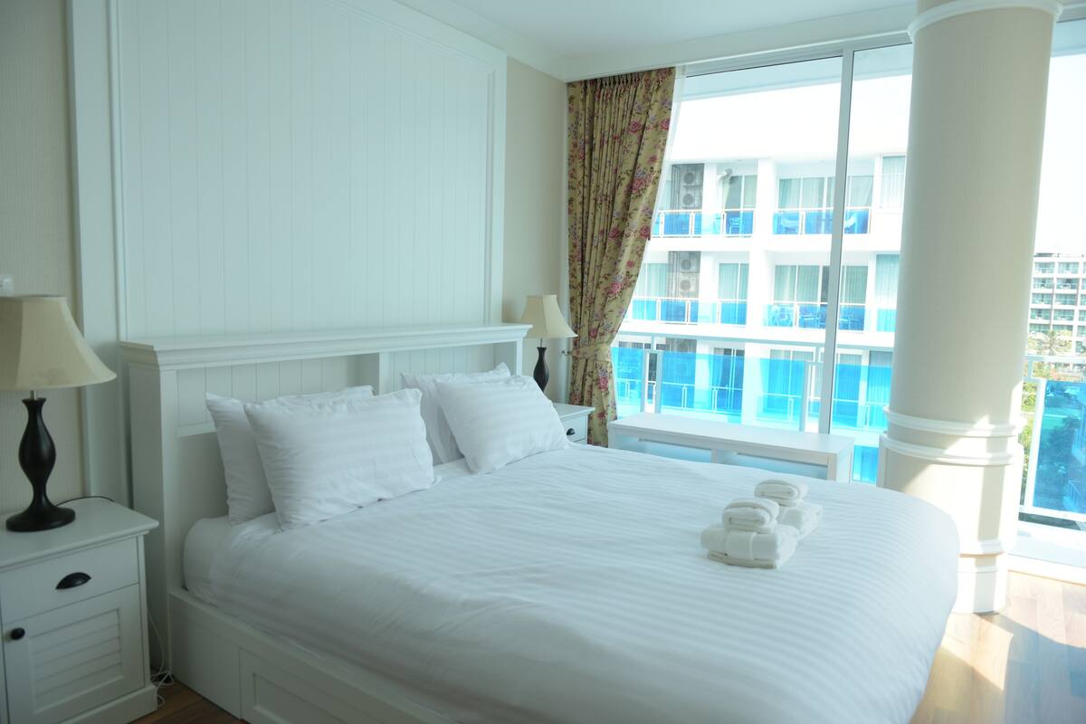 Une chambre d’hôtel avec un grand lit, une table de chevet avec une lampe, une fenêtre donnant sur un immeuble et un balcon avec une balustrade.
