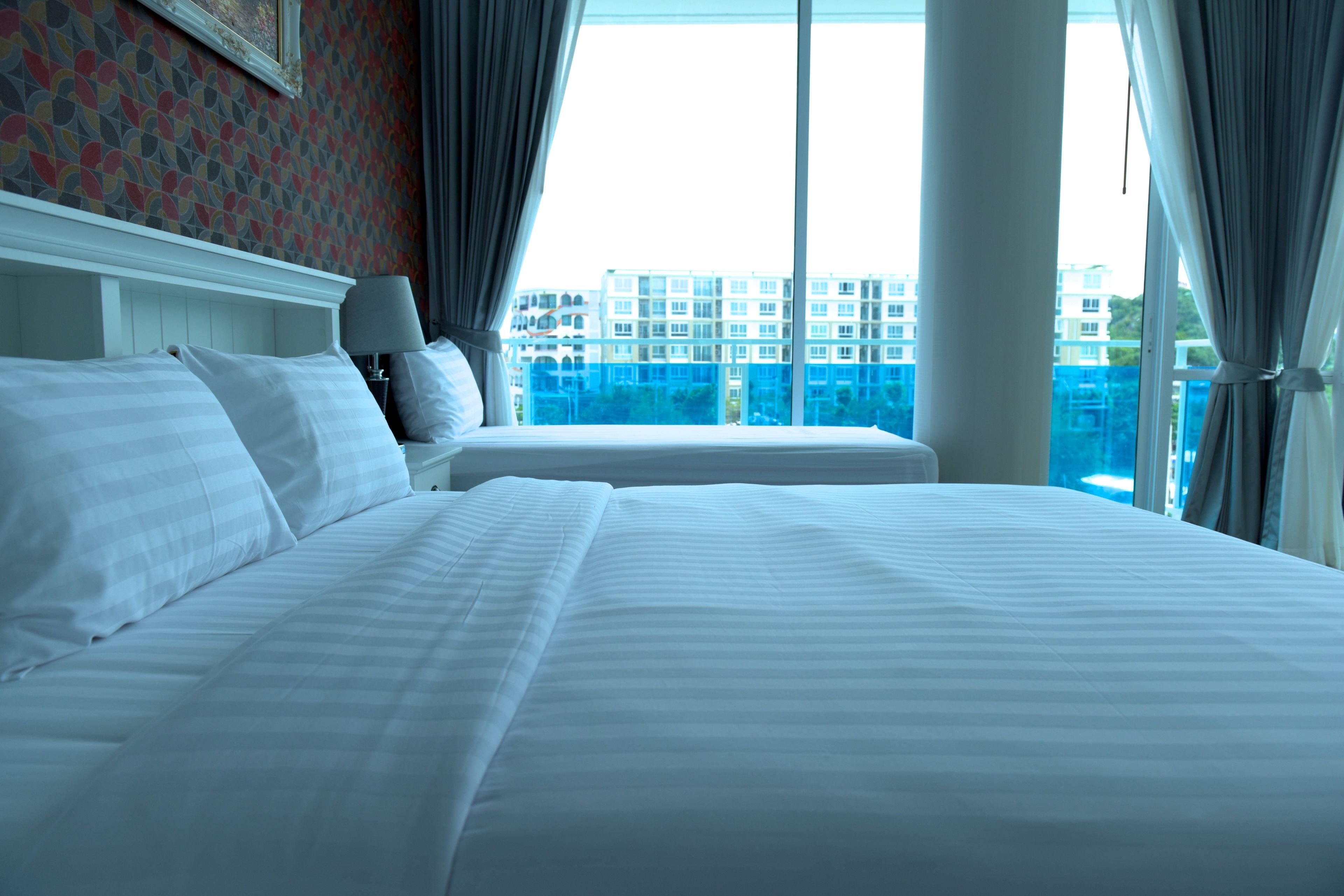 A hotel room with two beds, a large window with curtains, and a view of a blue building.