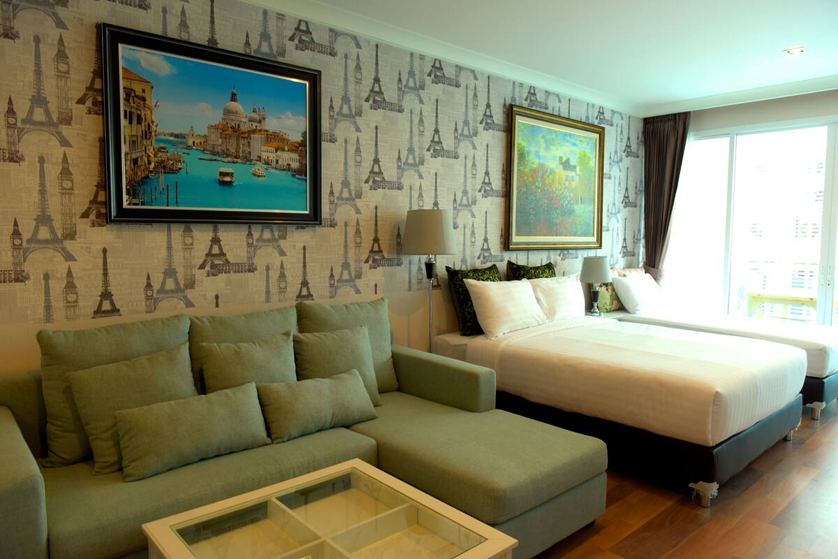 Une chambre d’hôtel avec un grand lit, un canapé et une table basse en verre. Les murs sont recouverts d’un papier peint orné d’un motif représentant la Tour Eiffel.