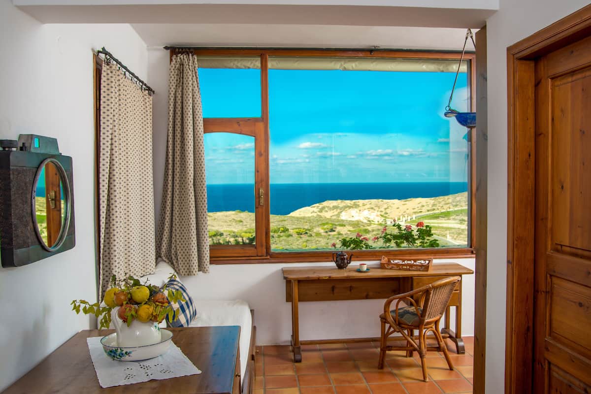 A room with a large window overlooking a coastal landscape, a wooden desk with a chair, and a vase of flowers on the windowsill.