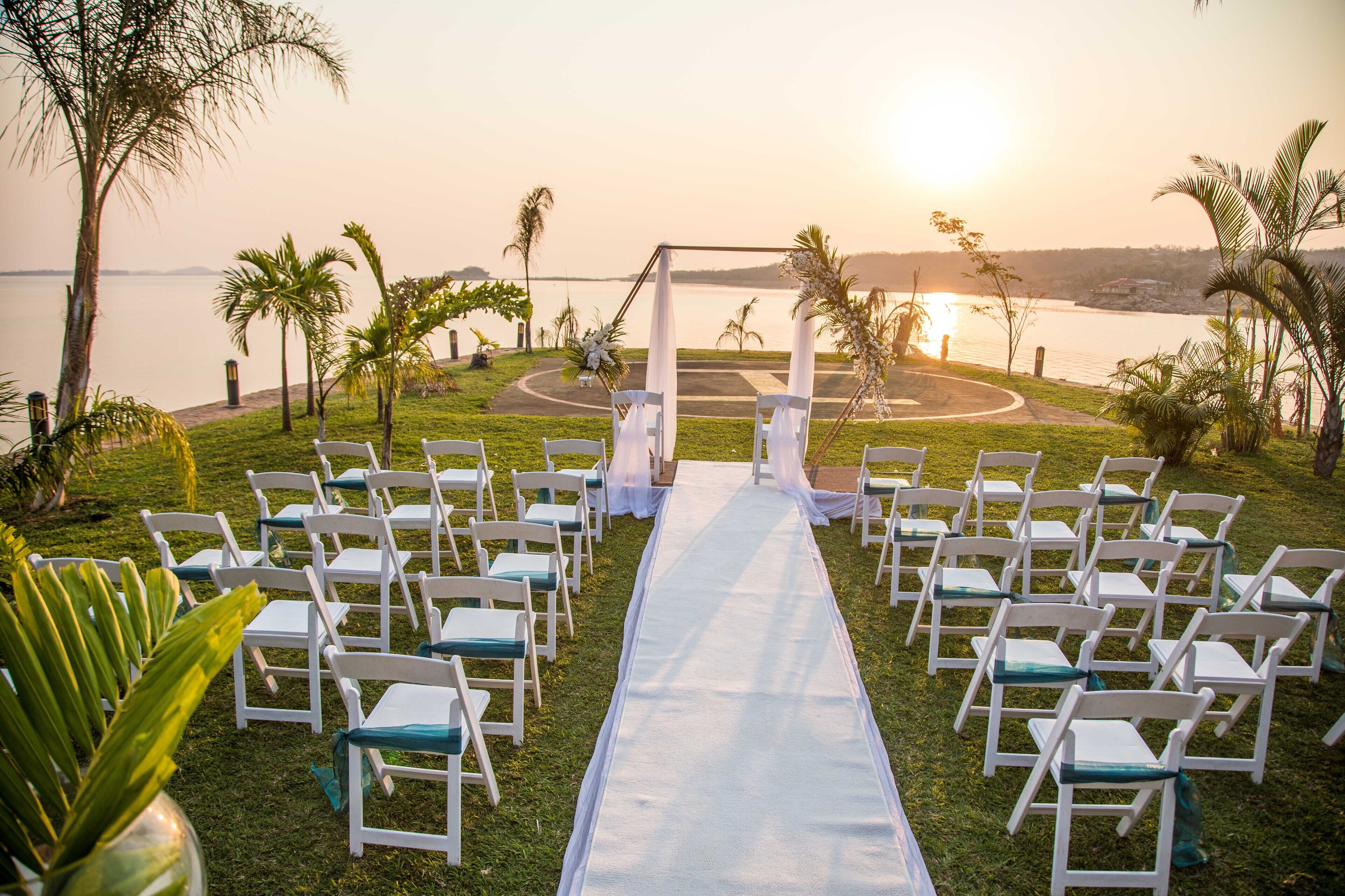 An outdoor wedding setup with a white aisle, white chairs, and a backdrop of palm trees and a body of water.