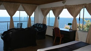 Een kamer met een bed, een leren bank, een bureau en een stoel. De kamer heeft grote ramen met gordijnen en uitzicht op de zee.