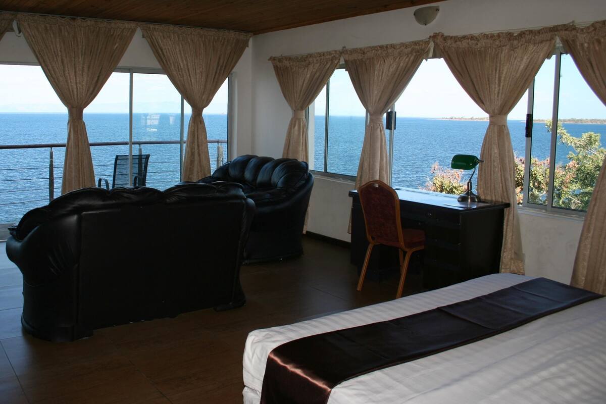 A room with a bed, a leather sofa, a desk, and a chair. The room has large windows with curtains, offering a view of the sea.