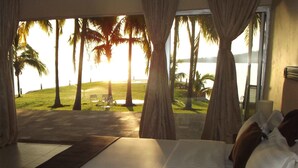 Een slaapkamer met een groot bed, gordijnen en uitzicht op palmbomen en een strand.