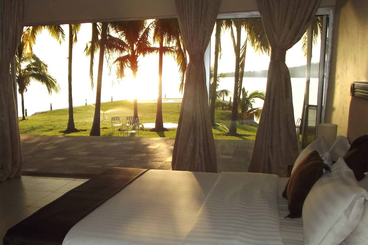 A bedroom with a large bed, curtains, and a view of palm trees and a beach.