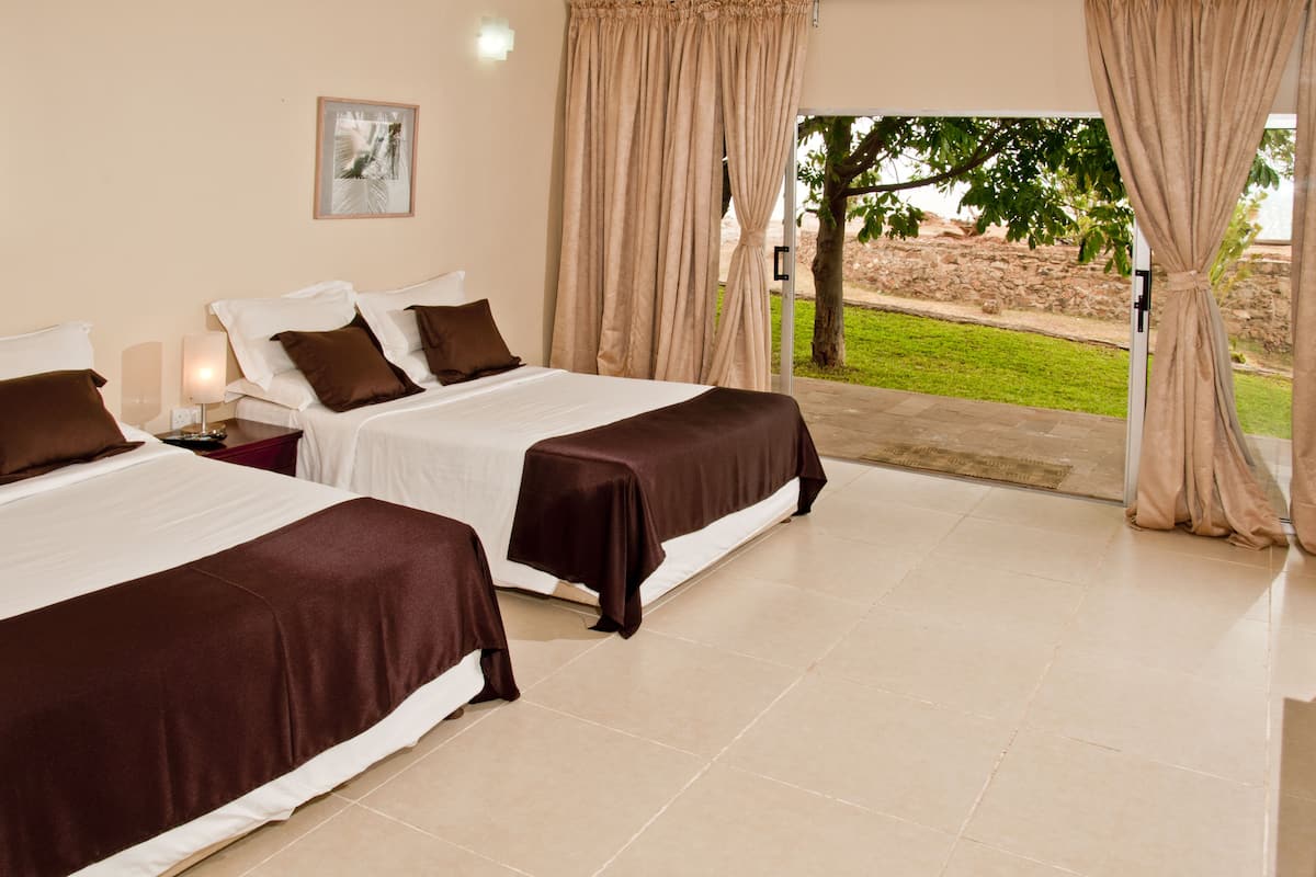 A hotel room with two beds, a nightstand, a lamp, and a view of the outdoors.