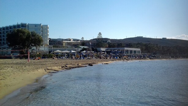 A beach with clear water, a sandy shore, and buildings in the background. - Anna Hotel (Hersonissos)