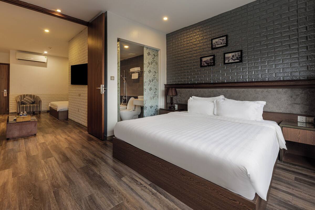 A hotel room with a large bed, a wooden headboard, a brick accent wall, and a small bedside table with a lamp.