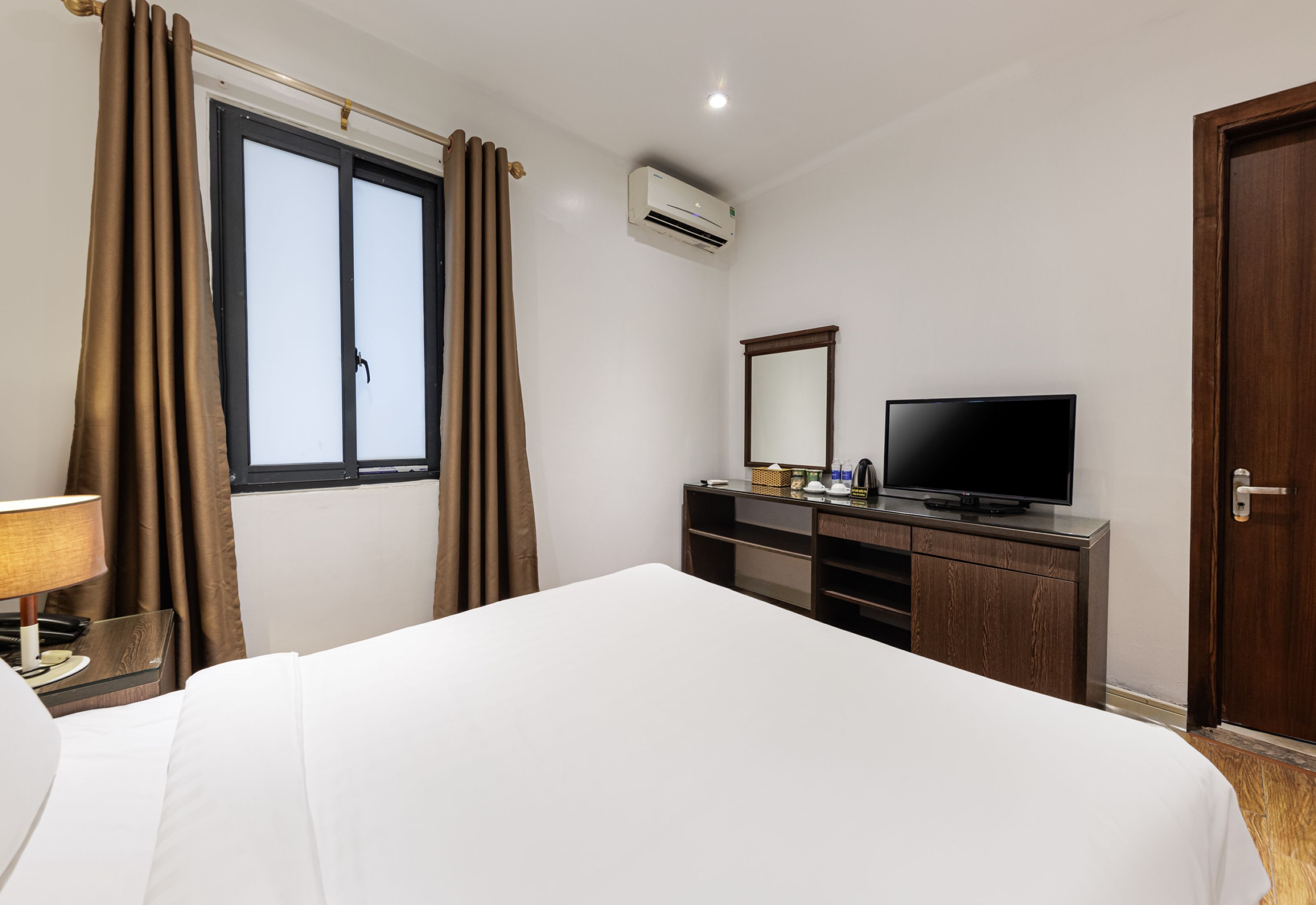 Een hotelkamer met een bed, een televisie op een houten dressoir, een raam met gordijnen, een airconditioner en een deur.