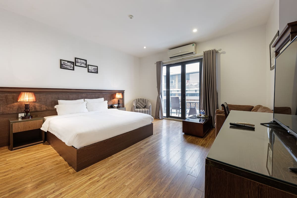 A hotel room with a large bed, wooden flooring, a desk with a TV, and a balcony with curtains.