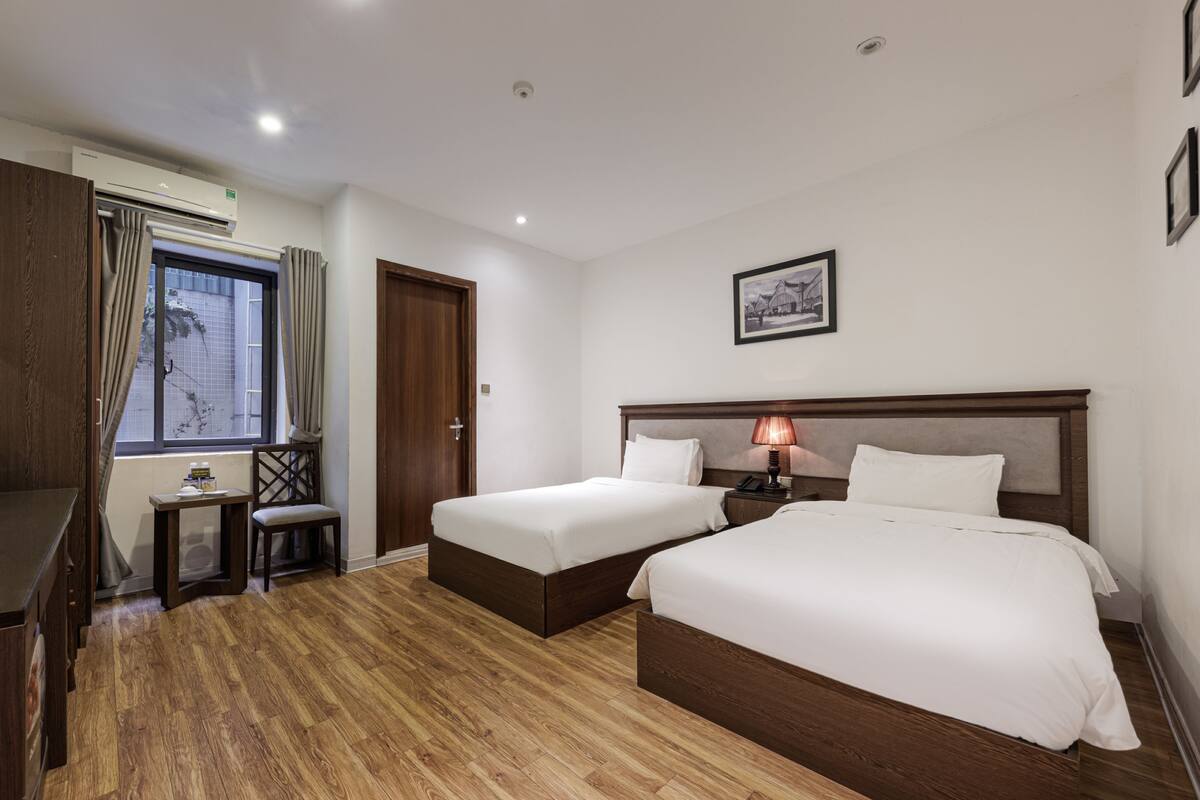 A hotel room with two beds, a desk, a chair, a window with curtains, and a framed picture on the wall.