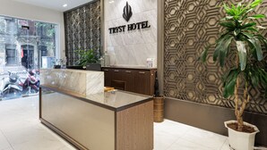 A reception desk with a marble top and wooden base, a sign reading "TRYST HOTEL", a decorative wall with geometric patterns, and a potted plant. - Collection O Le Grand Hanoi hotel – The Tryst (Hanoi)