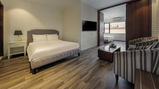 A hotel room with a large bed, a TV mounted on the wall, a seating area with striped chairs, and a wooden floor. - Collection O Le Grand Hanoi hotel – The Tryst (Hanoi)
