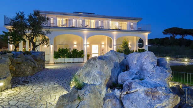 A two-story house with a balcony, stone pathway, and large rocks in the foreground. - Casa Colarusso (Massa Lubrense)