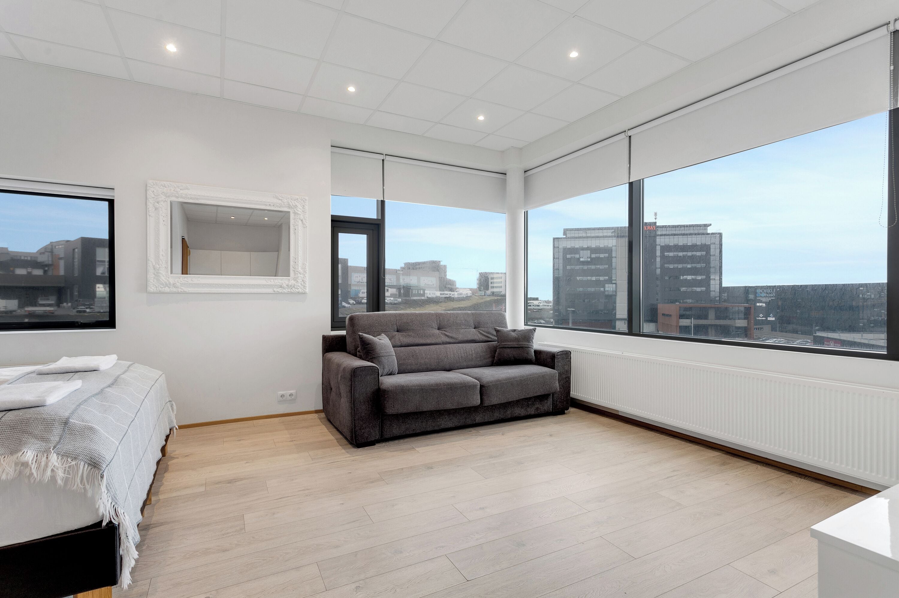 A modern bedroom with a large window, a grey sofa, a bed with a white and grey striped blanket, and a white radiator.