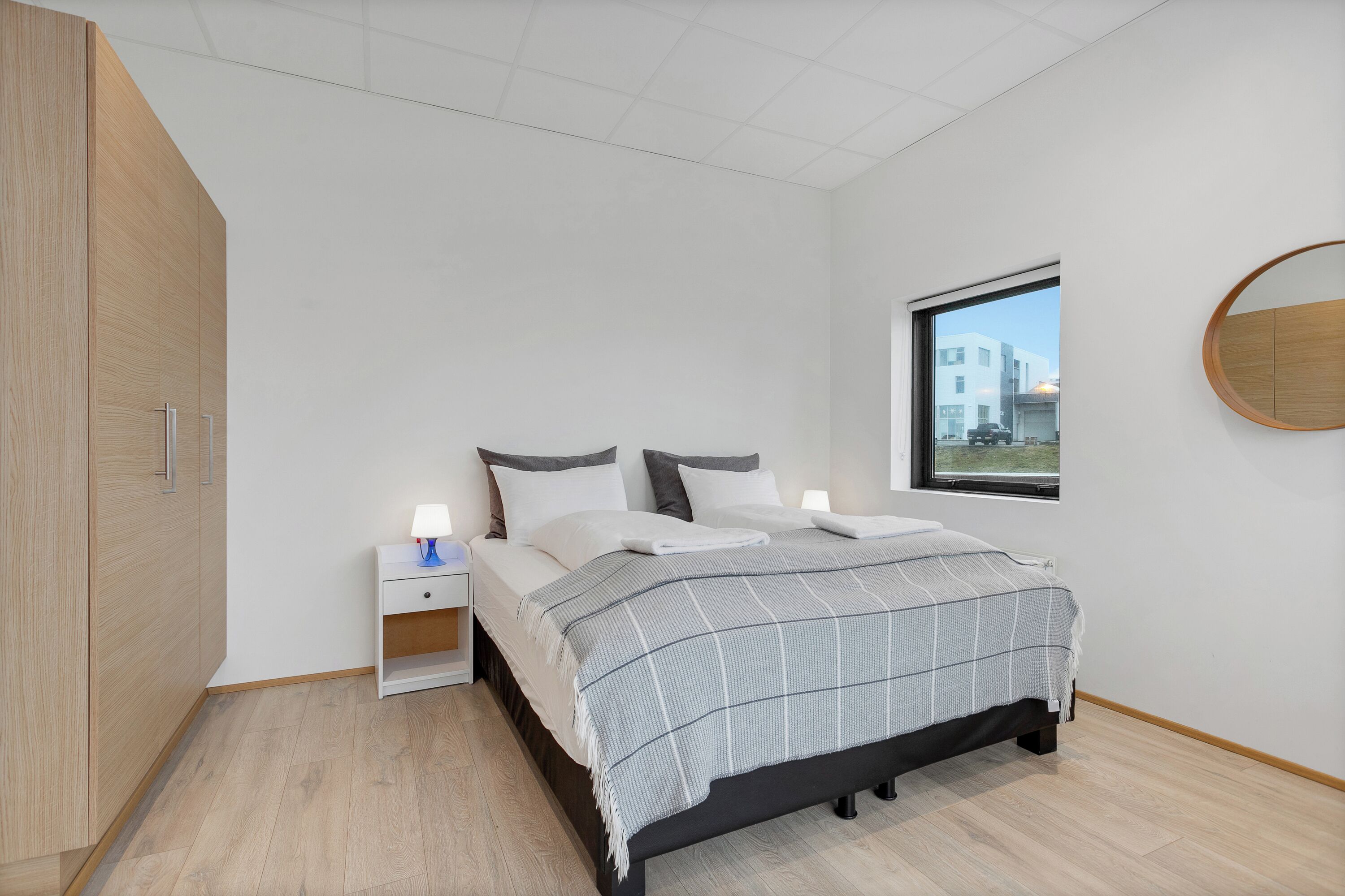 A bedroom with a bed, bedside table, lamp, and a window with a view of buildings.