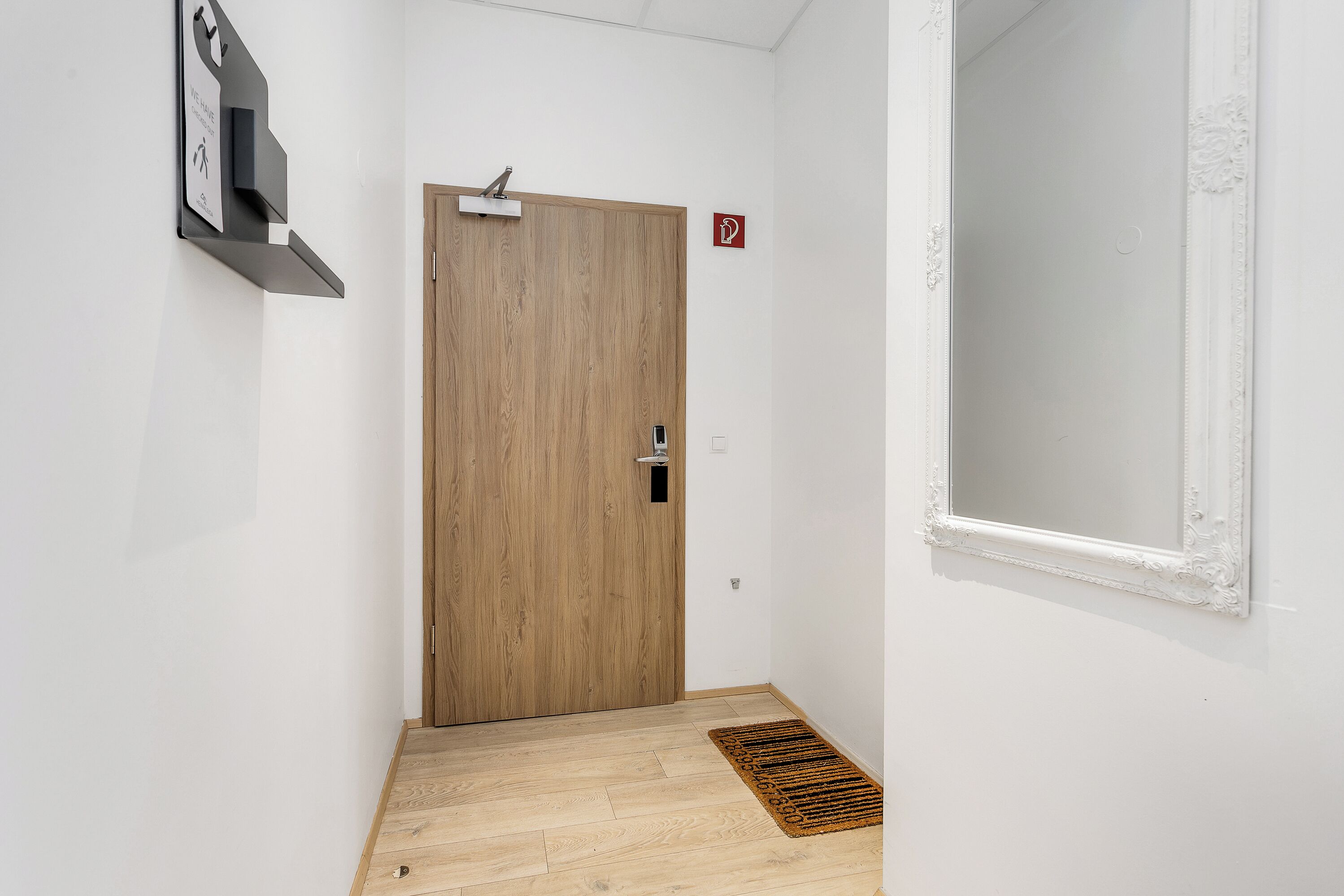 A narrow hallway with a wooden door, a small shelf with a sign, and a window with a white frame.