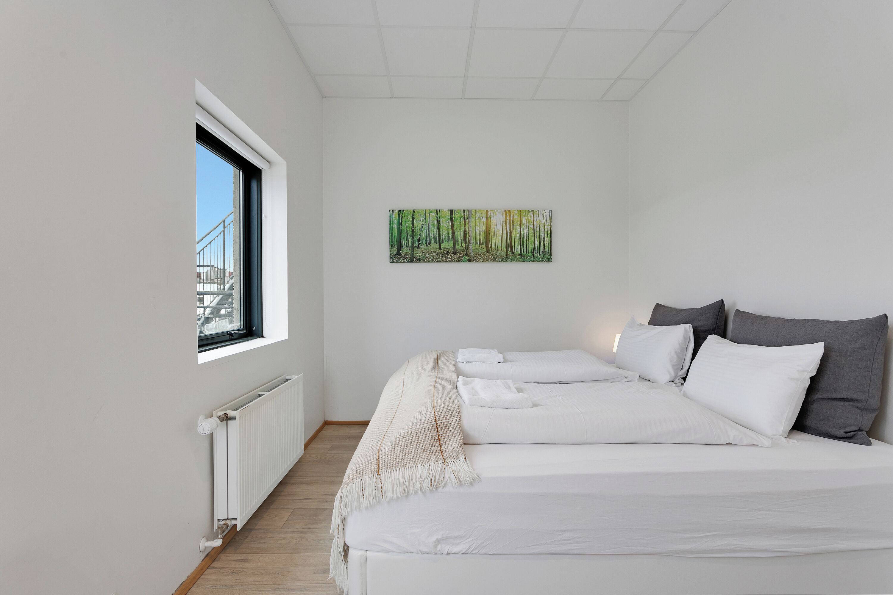 Ein modernes Hotelzimmer mit großem Fenster, einem Bett mit weißen Leinentüchern, einem Waldgemälde und einem Heizkörper an der Wand.