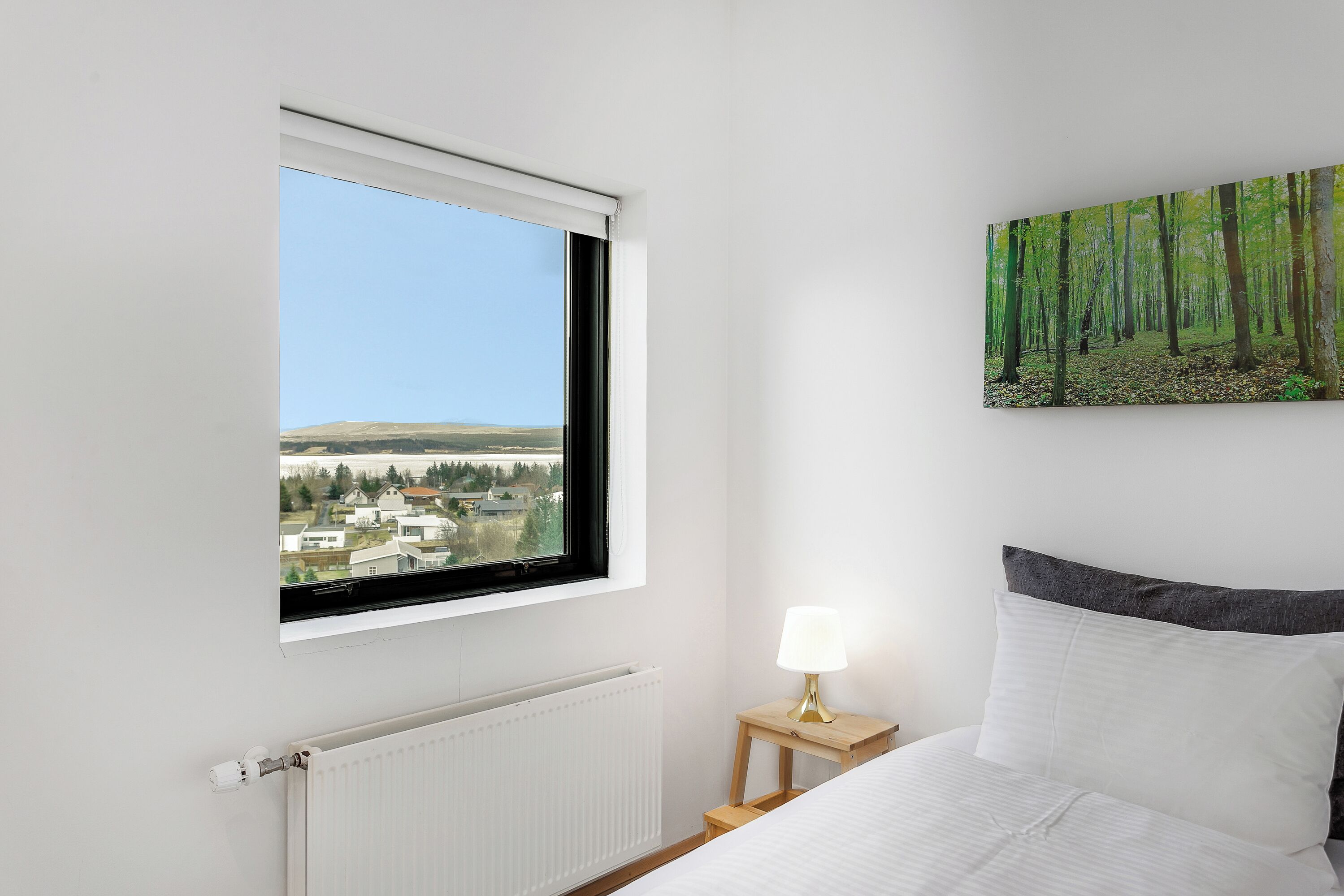 A bedroom with a window, a painting of a forest, a bedside lamp, and a bed with white bedding.