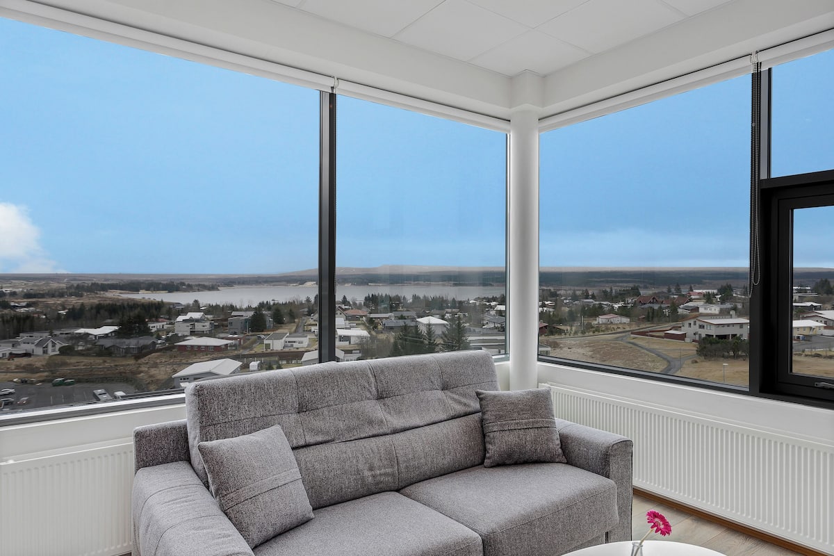 A modern living room with a grey sofa, large windows offering a view of a town and water body.