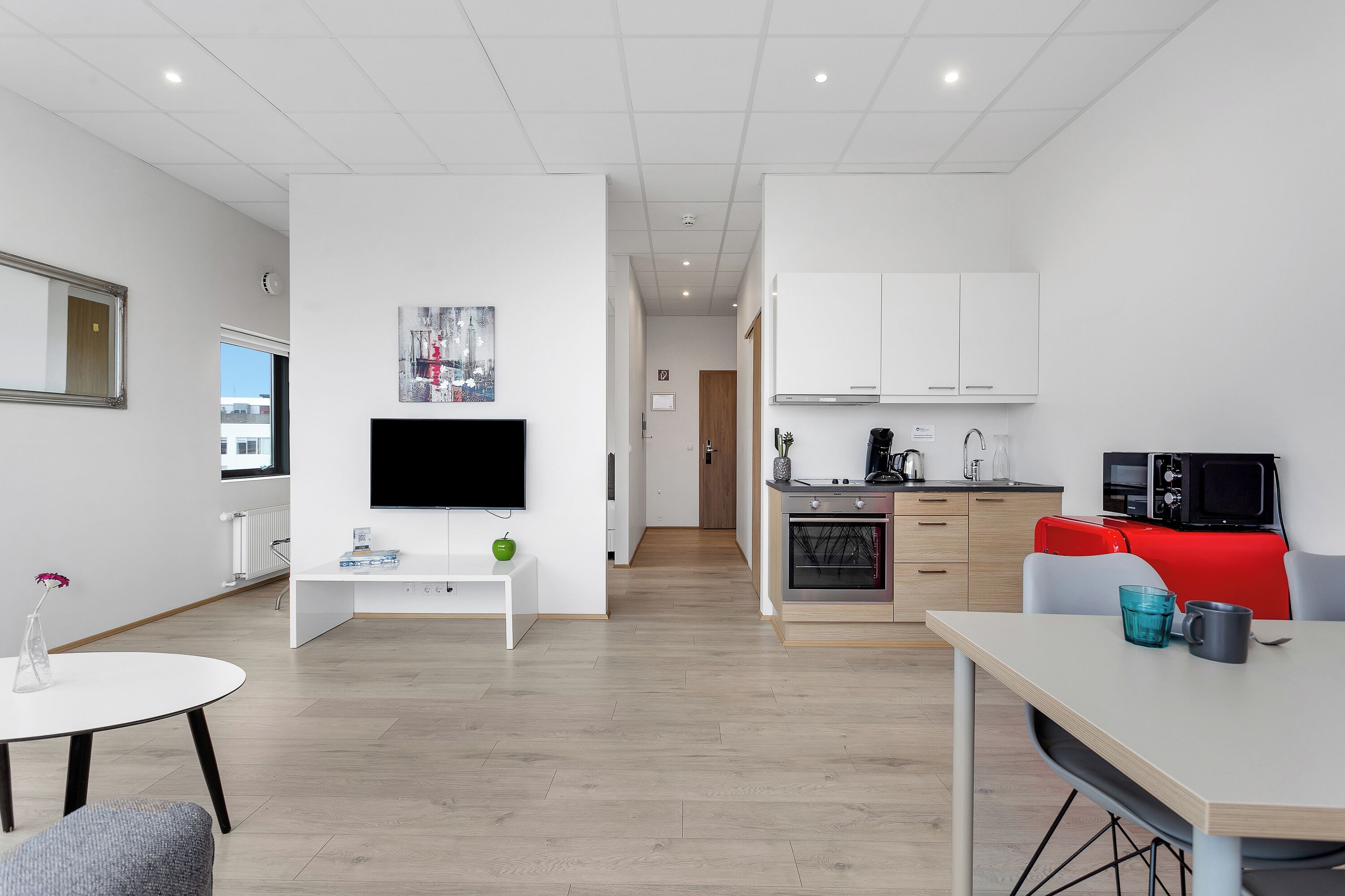 A modern living room with a flat-screen TV, a small white table, and a red couch. There is a kitchen area with white cabinets and a microwave.
