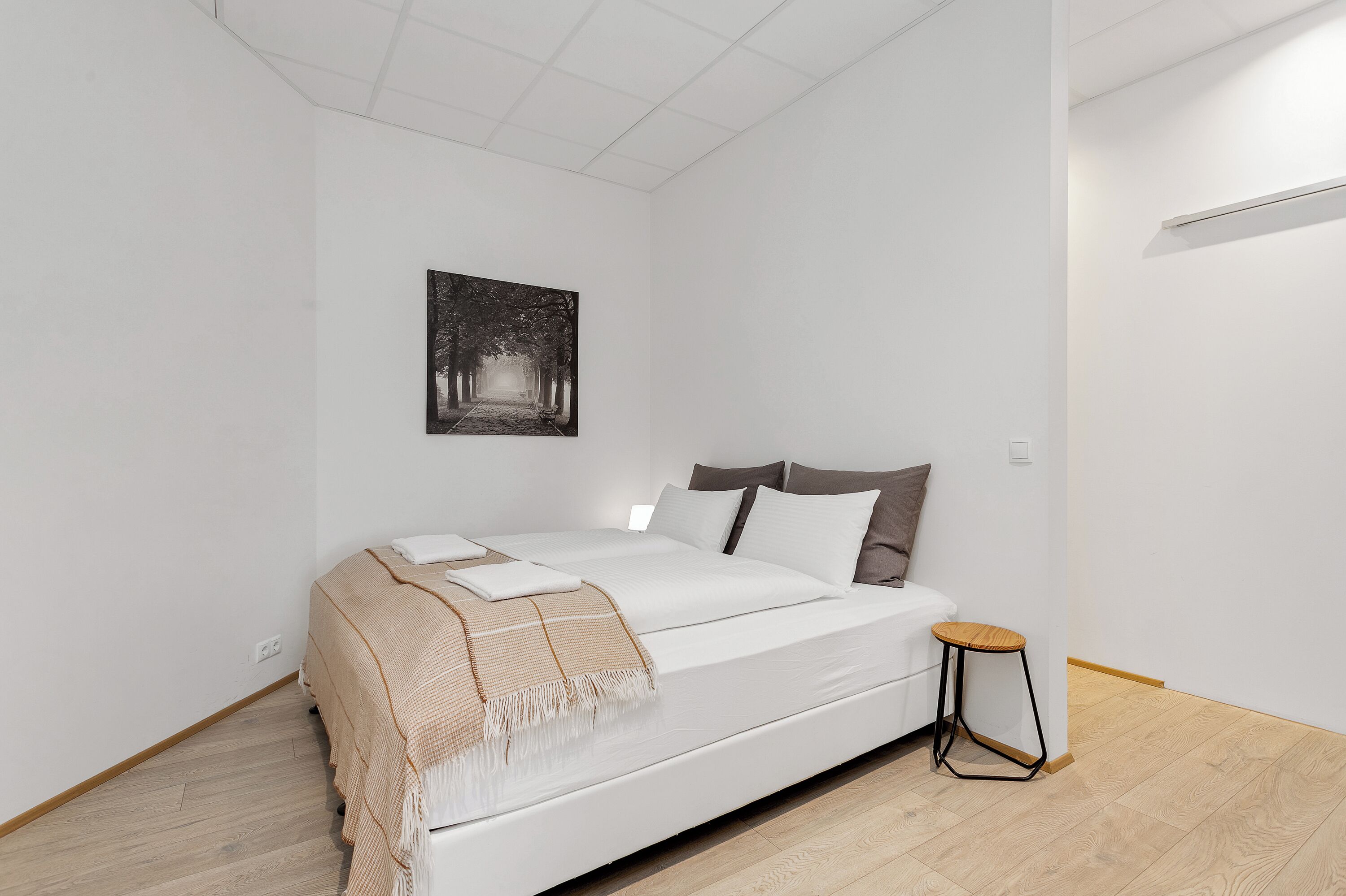 Een minimalistische slaapkamer met een groot bed, een klein krukje en een foto aan de muur.