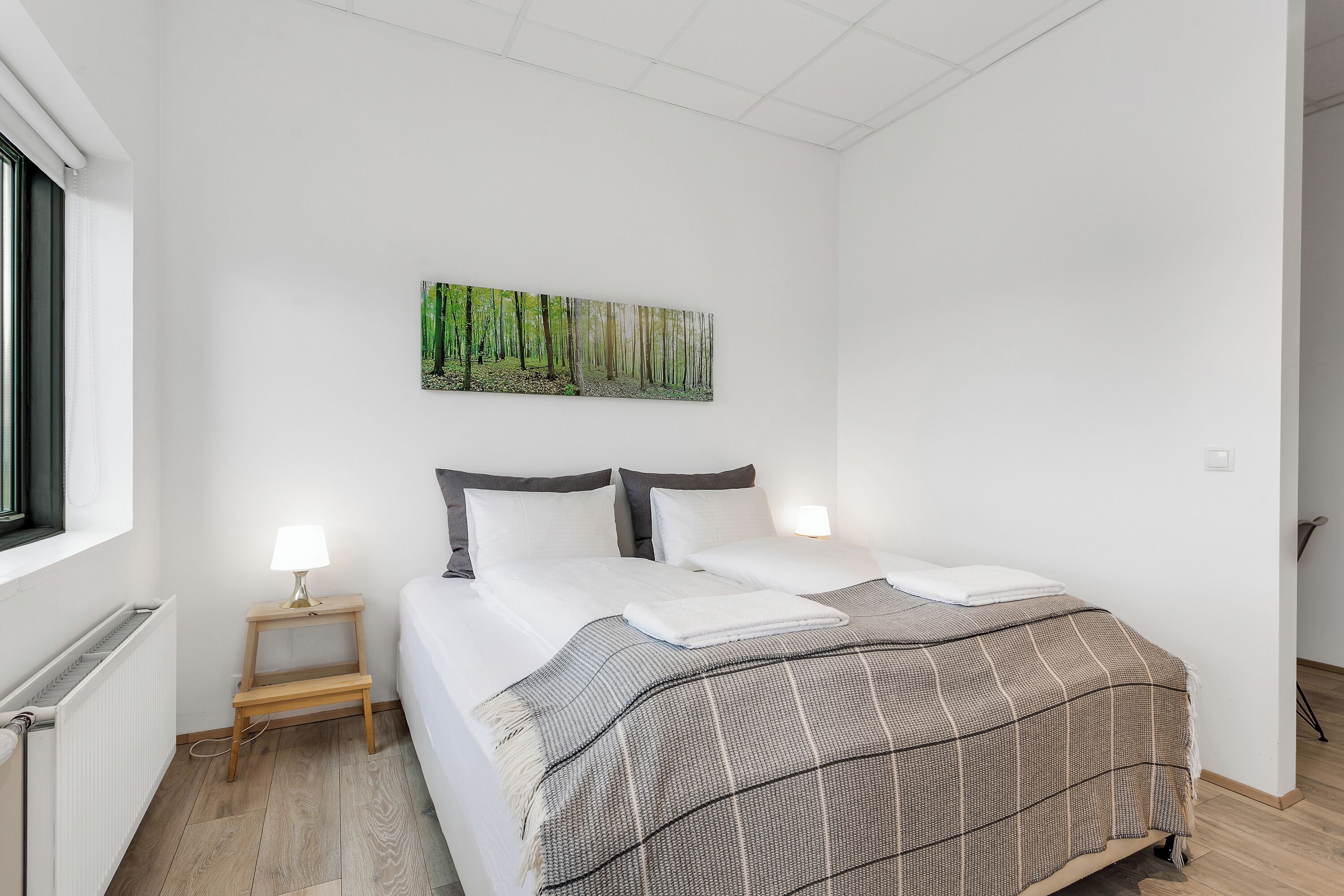 Una camera da letto con un letto, due lampade da comodino, un piccolo sgabello di legno, un termosifone e un quadro raffigurante una foresta appeso al muro.