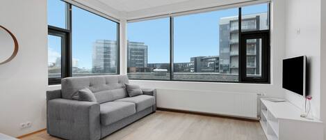 Ein modernes Wohnzimmer mit grauem Sofa, großen Fenstern, einem weißen TV-Schrank und Blick auf die Stadtlandschaft.