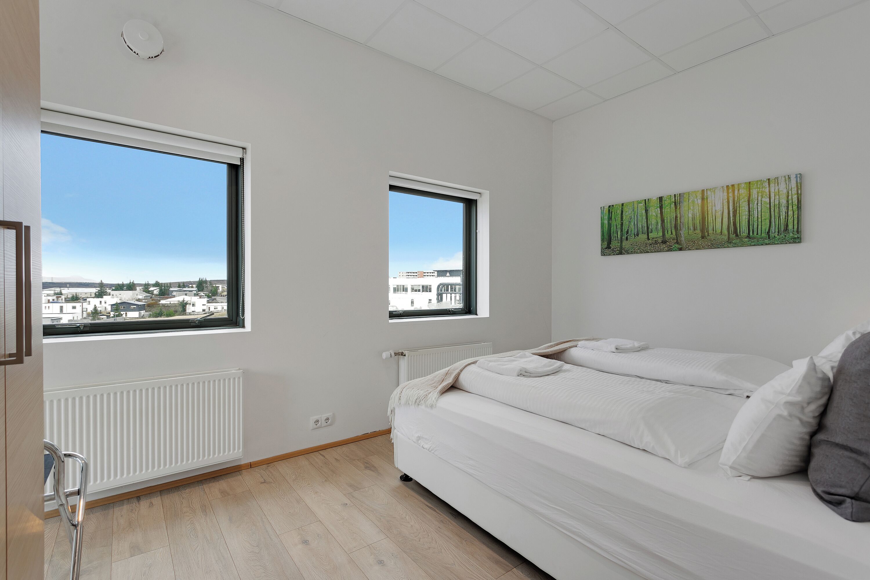 A bedroom with a large bed, two windows, a radiator, and a view of buildings.