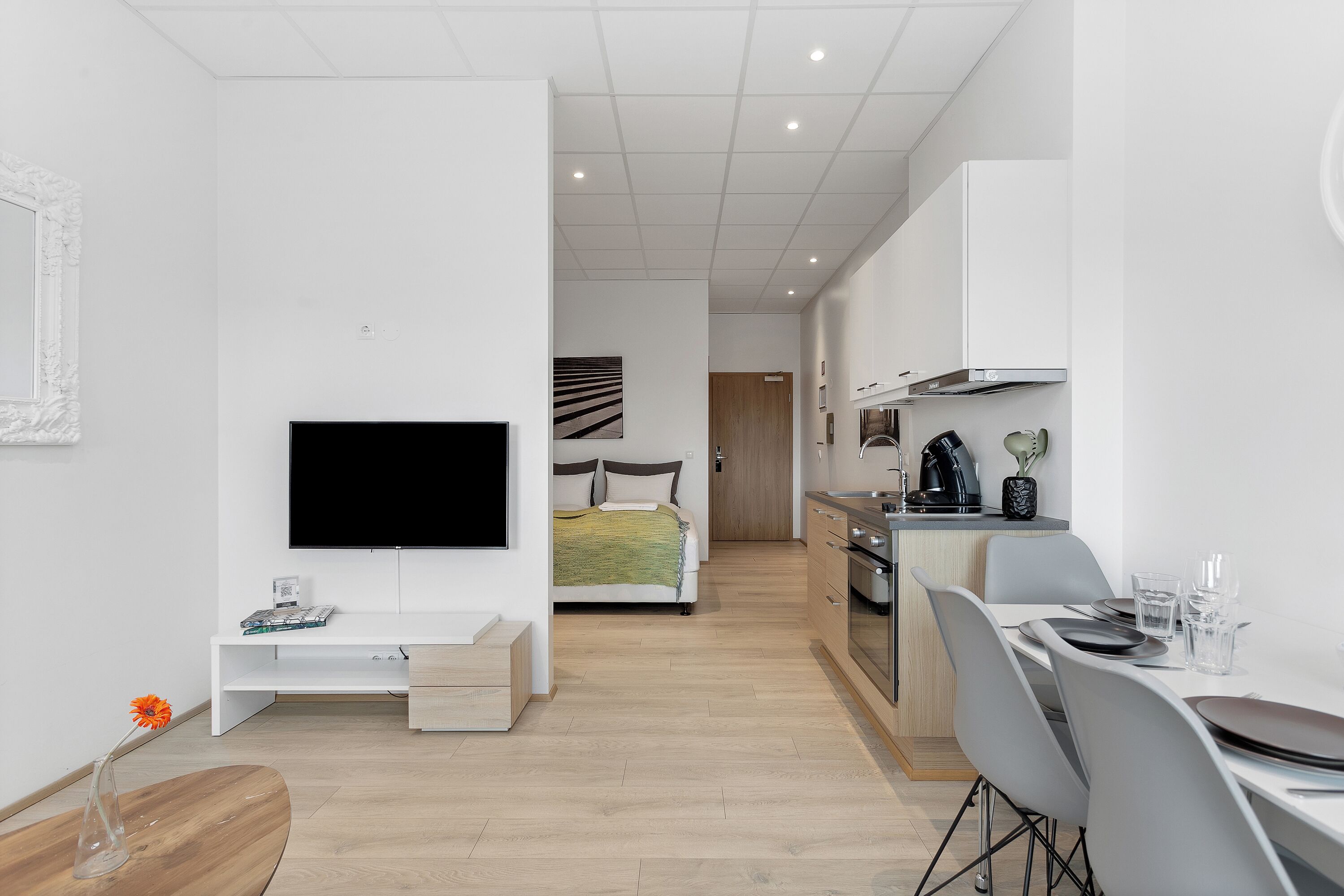 A modern, well-lit apartment with a living area featuring a flat-screen TV, a dining table with chairs, a kitchen with a stove and sink, and a bedroom with a bed and a large window.