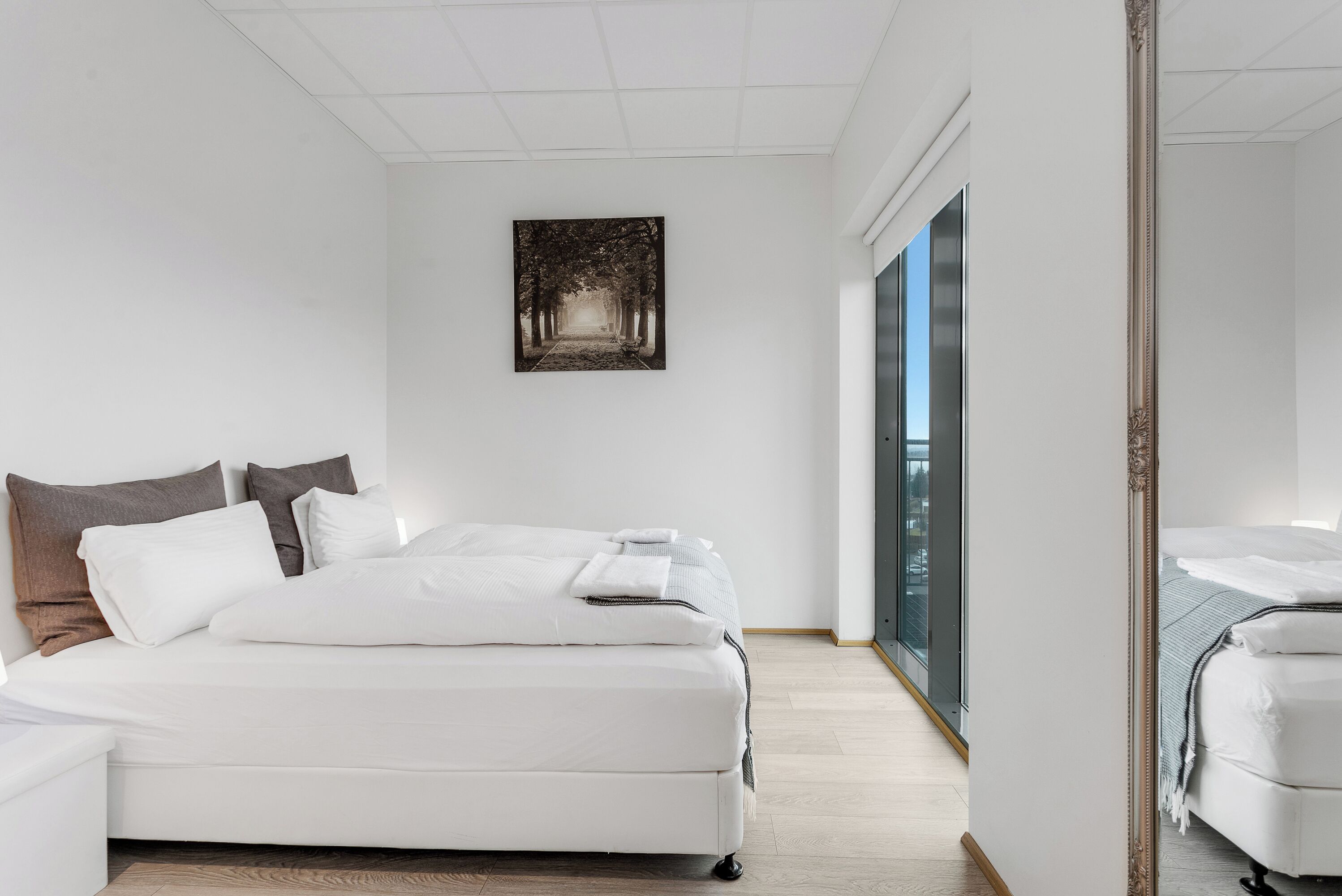 A modern hotel room with a large bed, a bedside table, a window with a view, and a framed artwork on the wall.