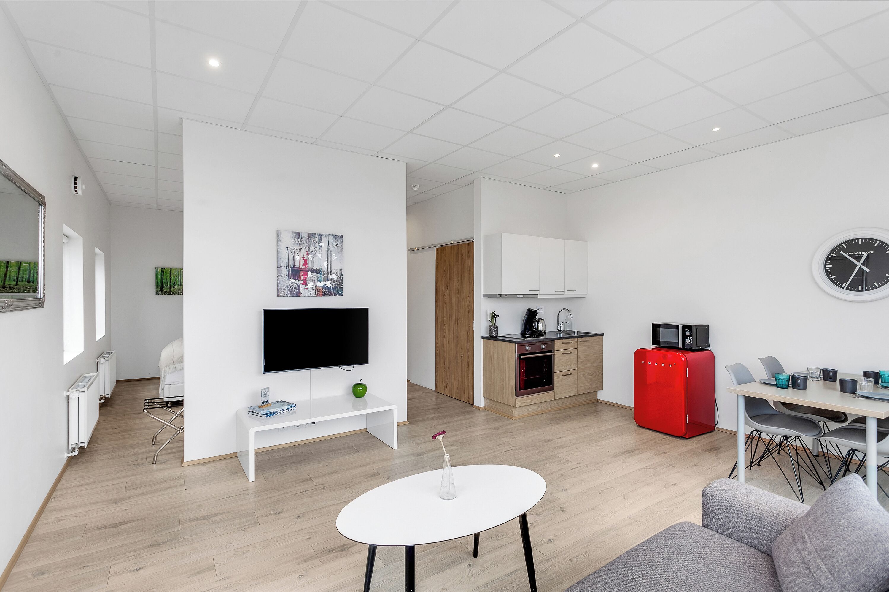Un salon moderne et bien éclairé, doté d’un téléviseur à écran plat, d’une petite table blanche et d’un réfrigérateur rouge.