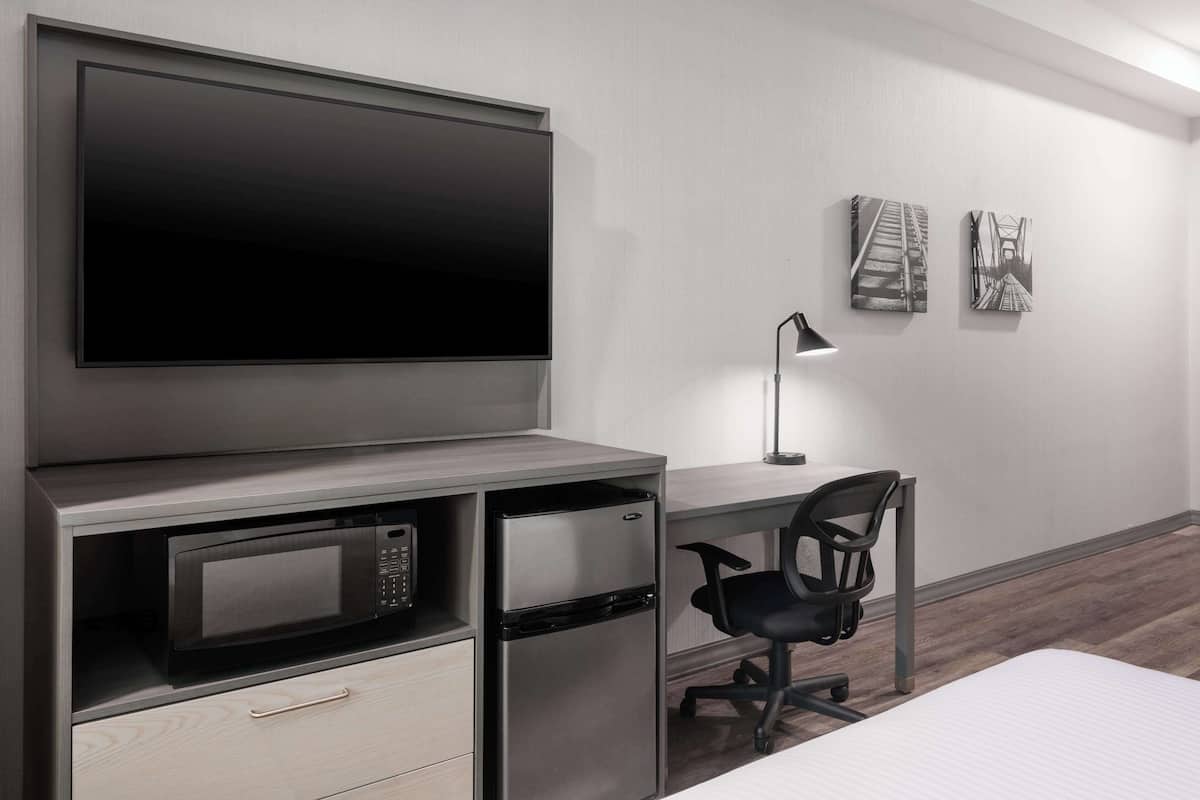 Ein modernes Zimmer mit einem Flachbildfernseher, der über einem Holztisch angebracht ist. Darunter befinden sich eine Mikrowelle und ein Kühlschrank. Ein schwarzer Bürostuhl und eine Schreibtischlampe sind ebenfalls vorhanden. An der Wand hängen zwei gerahmte Bilder.