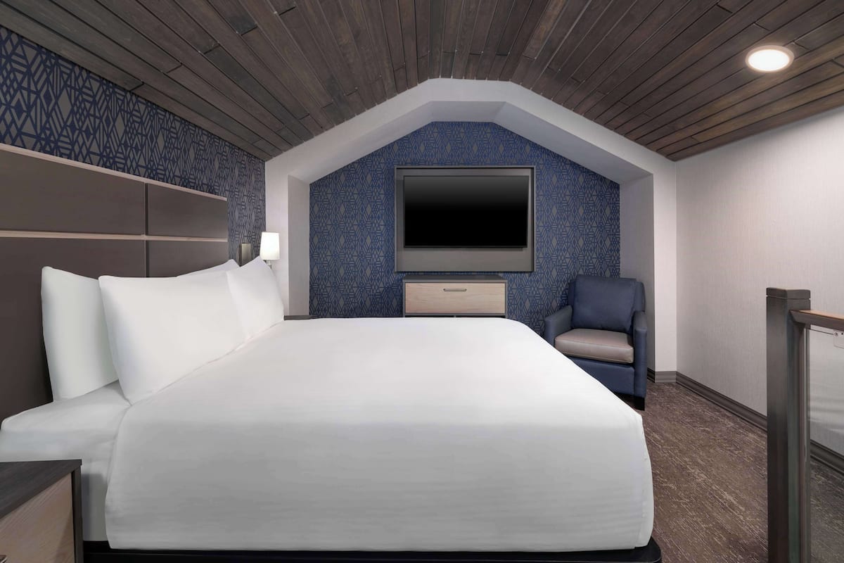 Ein Hotelzimmer mit einem großen Bett, einem flachbildfernseher an der Wand, einem Sessel und einem kleinen Tisch.
