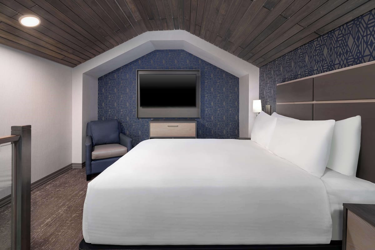 Ein Hotelzimmer mit einem großen Bett, einem an der Wand befestigten Fernseher, einem Sessel und einem Nachttisch.