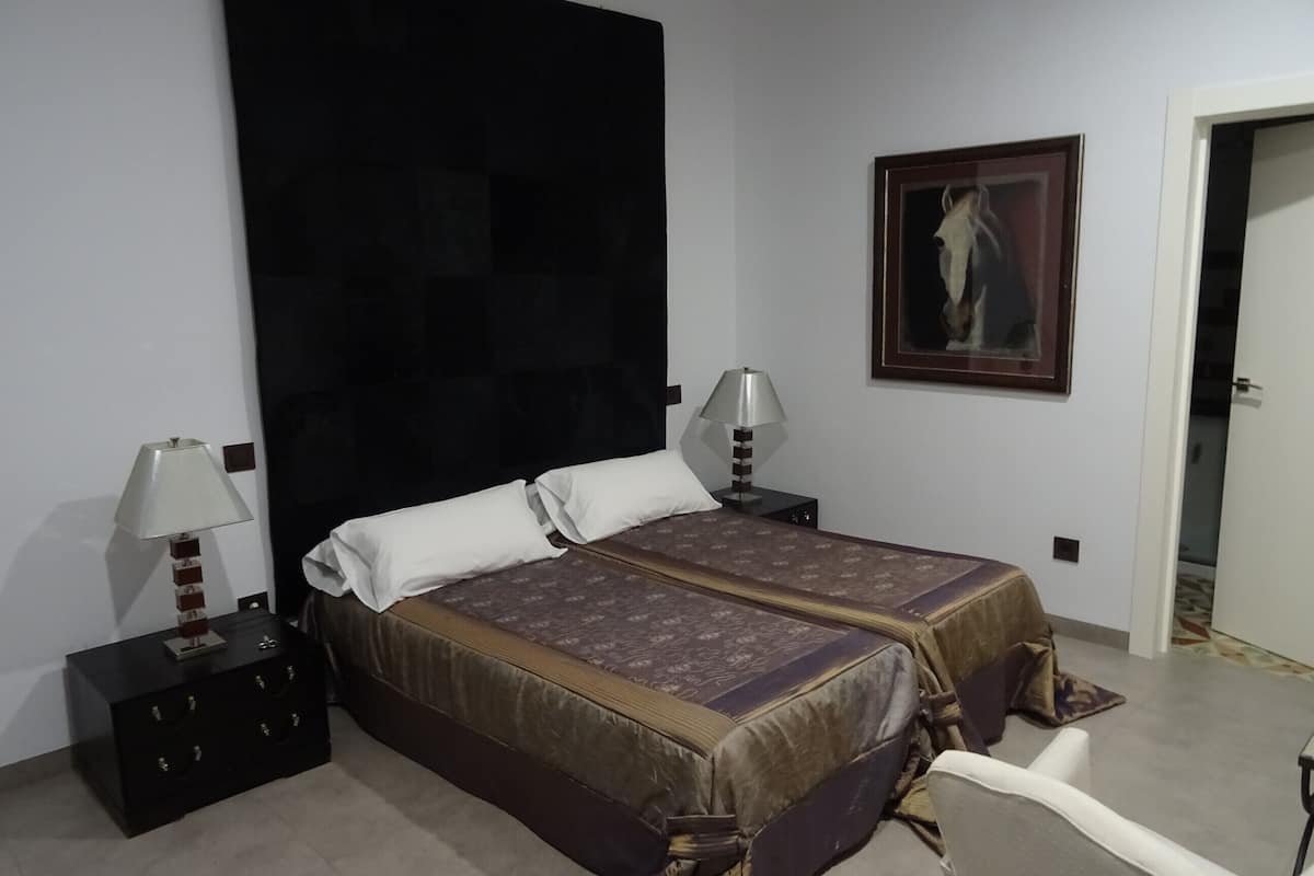 Quarto com duas camas, uma mesa de cabeceira com abajur, um quadro na parede e uma porta que dá para outro cômodo.