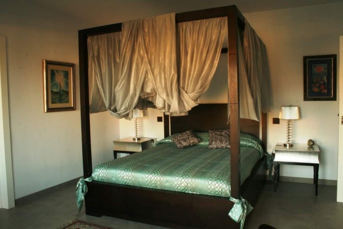 Uma cama de dossel com colcha verde estampada, duas mesas de cabeceira com abajures e um quadro na parede.