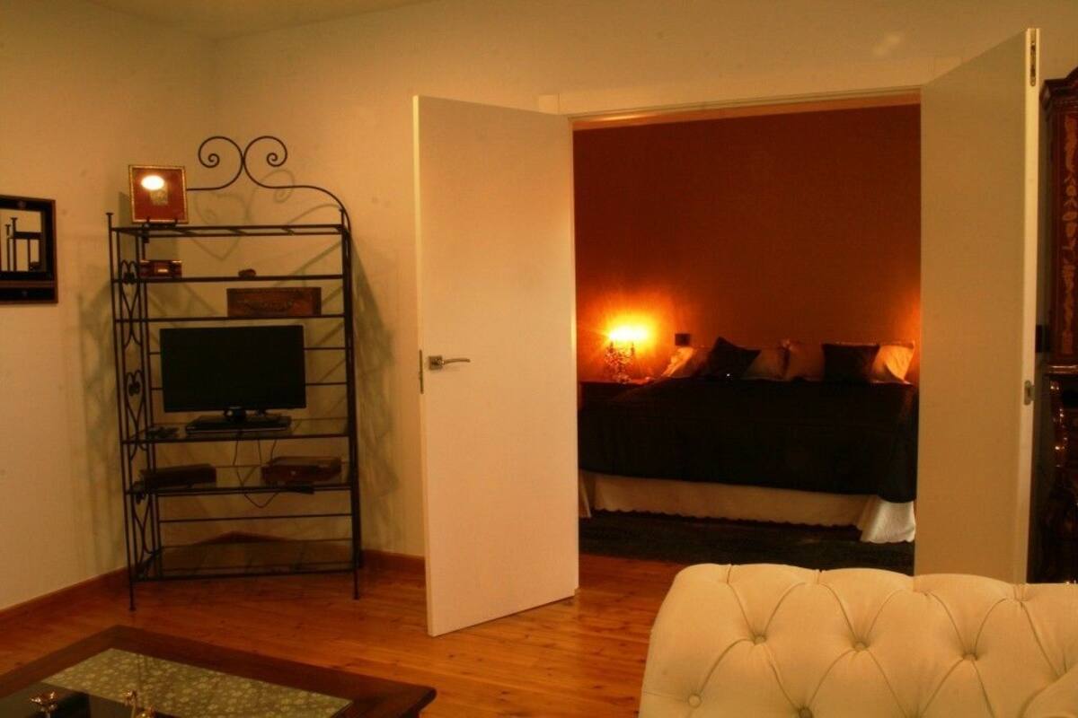 Quarto com televisão sobre suporte metálico, porta que dá para outro cômodo e cama com manta.