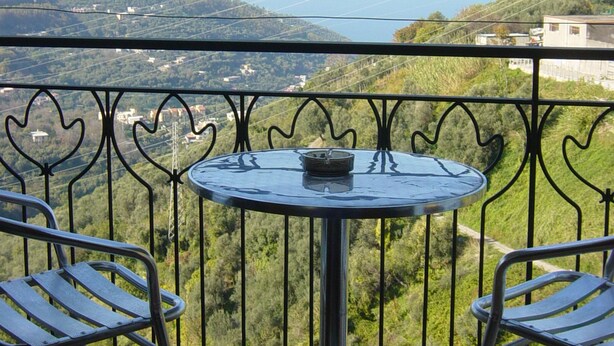 Un balcón con una mesa redonda y dos sillas, con vistas a un paisaje montañoso y al mar.