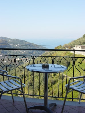 A balcony with a round table and two chairs, overlooking a hilly landscape with a view of the sea. - B&B Puntaferano (Vico Equense)