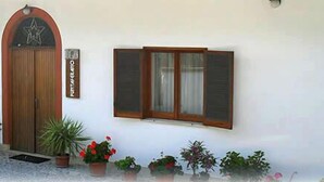 Un edificio con puerta de madera, ventana con contraventanas y varios maceteros con plantas al frente.