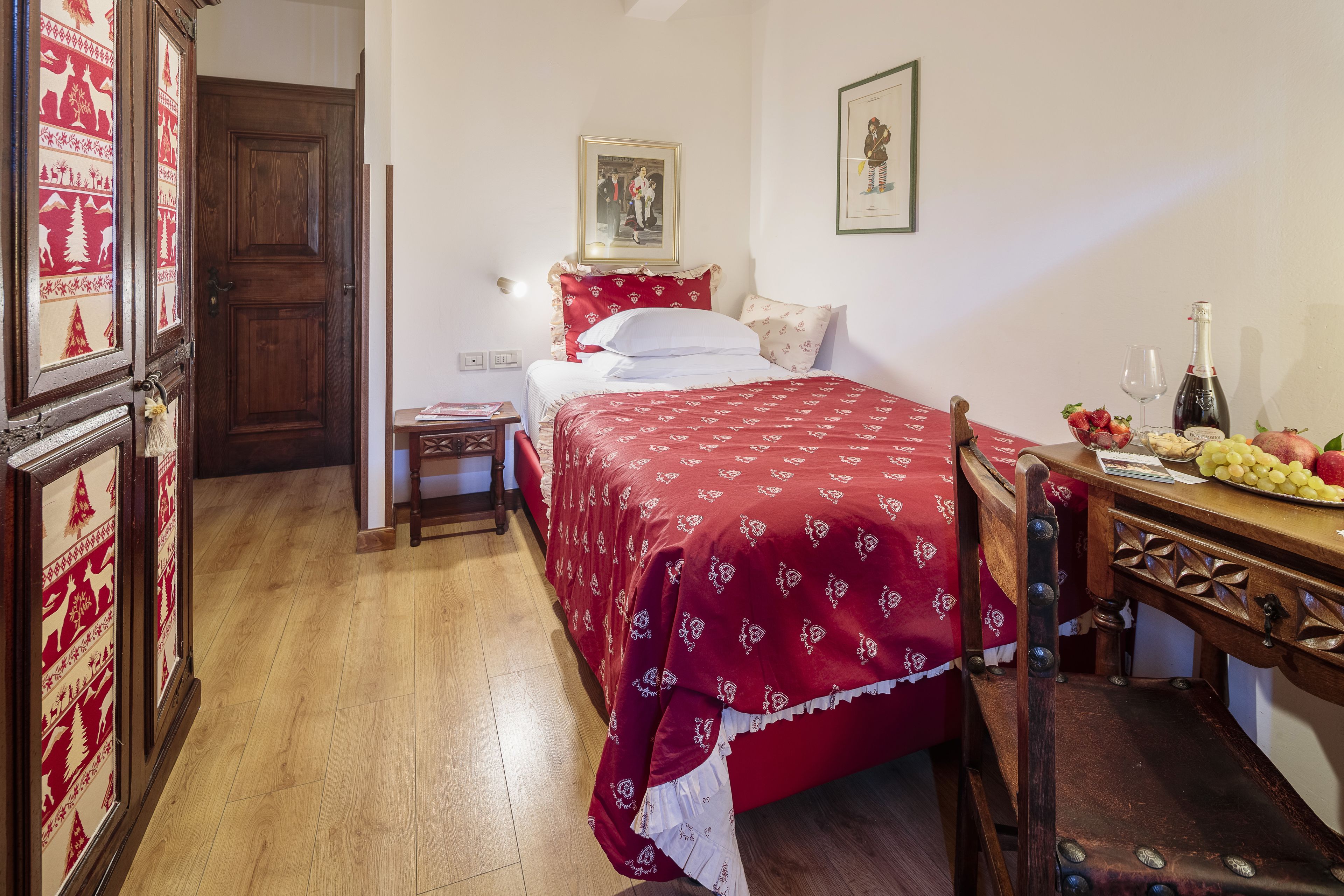 Un dormitorio con suelo de madera, una cama con manta roja, una silla de madera, una mesita con una botella y fruta, y una puerta con diseño rojo y blanco.