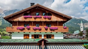 Un edificio de madera de dos pisos con balcones decorados con flores moradas, con montañas al fondo.