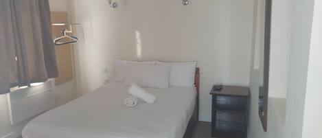 A neatly made bed with white linens, a small bedside table with a remote control, and a wall-mounted light fixture.